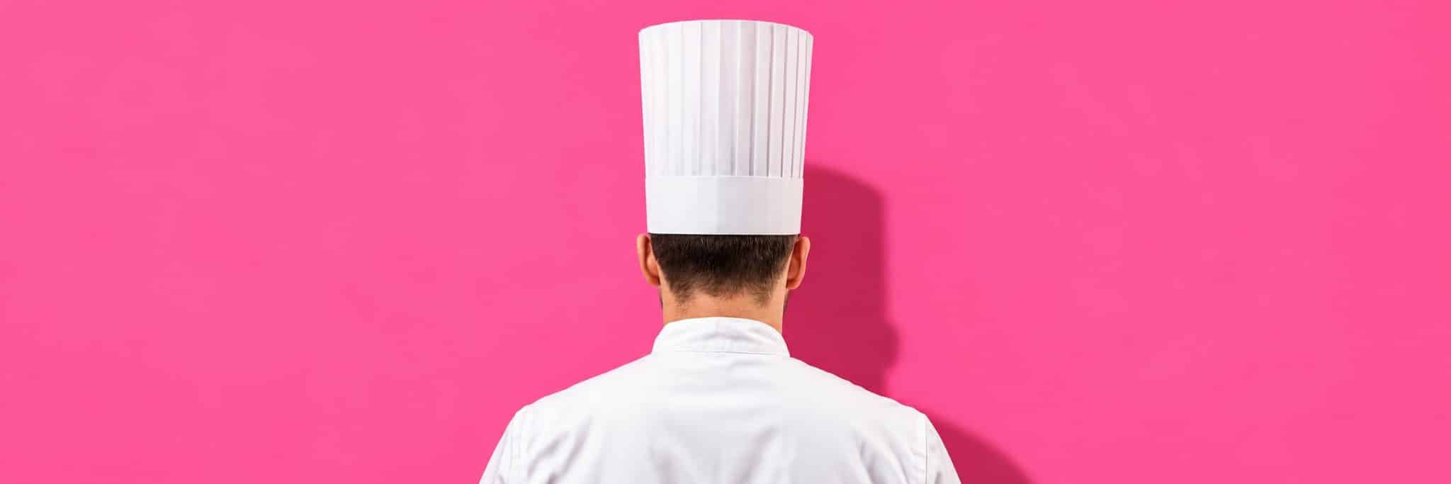 Cuisinier de dos, en veste blanche et toque haute, face à un mur rose