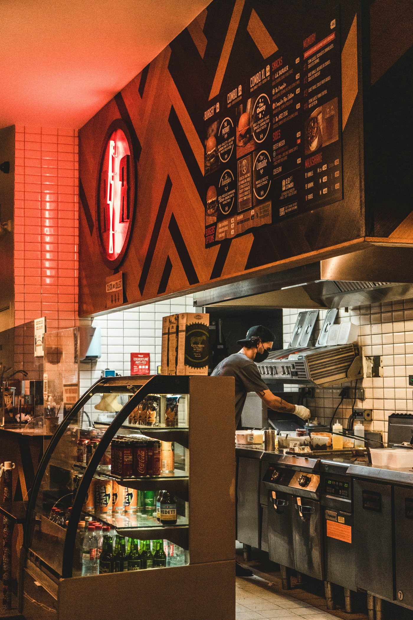 Cuisine ouverte d'un fast-food : un employé masqué et ganté prépare une commande derrière le comptoir, sous le menu lumineux